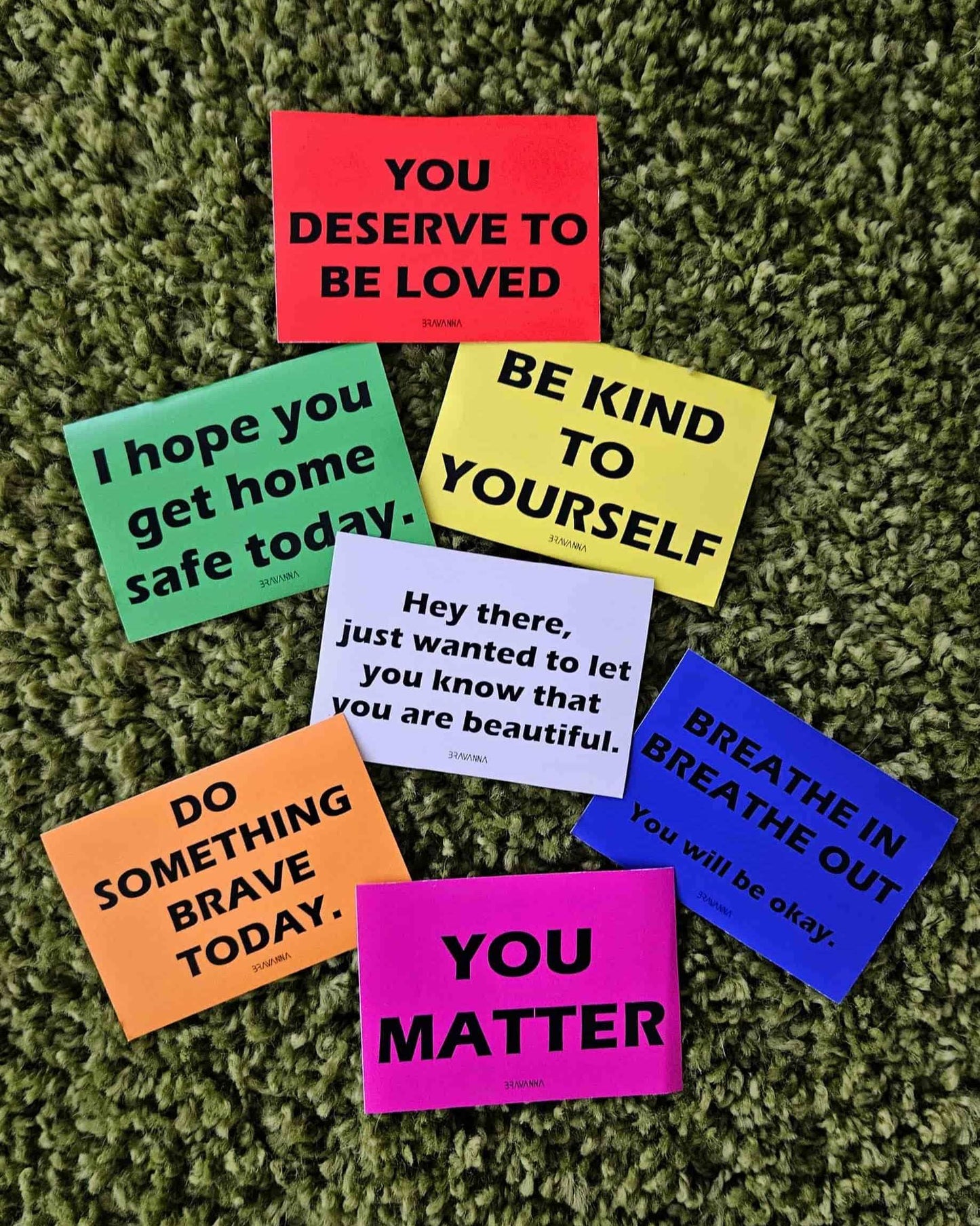 Green fabric textured carpet floor with seven colorful rectangular stickers laying on it. The colors are orange, pink, beige, red, yellow, blue and green. Messages on the stickers include each of these phrases: You matter, do something brave today, you deserve to be loved, be kind to yourself, breathe in breathe out you will be okay, I hope you get home safe today, hey there just wanted to let you know that you are beautiful.