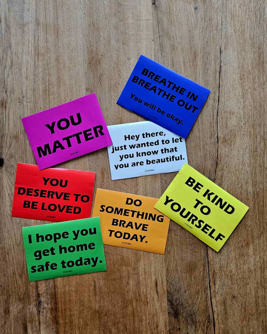 Wooden table with seven colorful rectangular stickers laying on it. The colors are orange, pink, beige, red, yellow, blue and green. Messages on the stickers include each of these phrases: You matter, do something brave today, you deserve to be loved, be kind to yourself, breathe in breathe out you will be okay, I hope you get home safe today, hey there just wanted to let you know that you are beautiful.