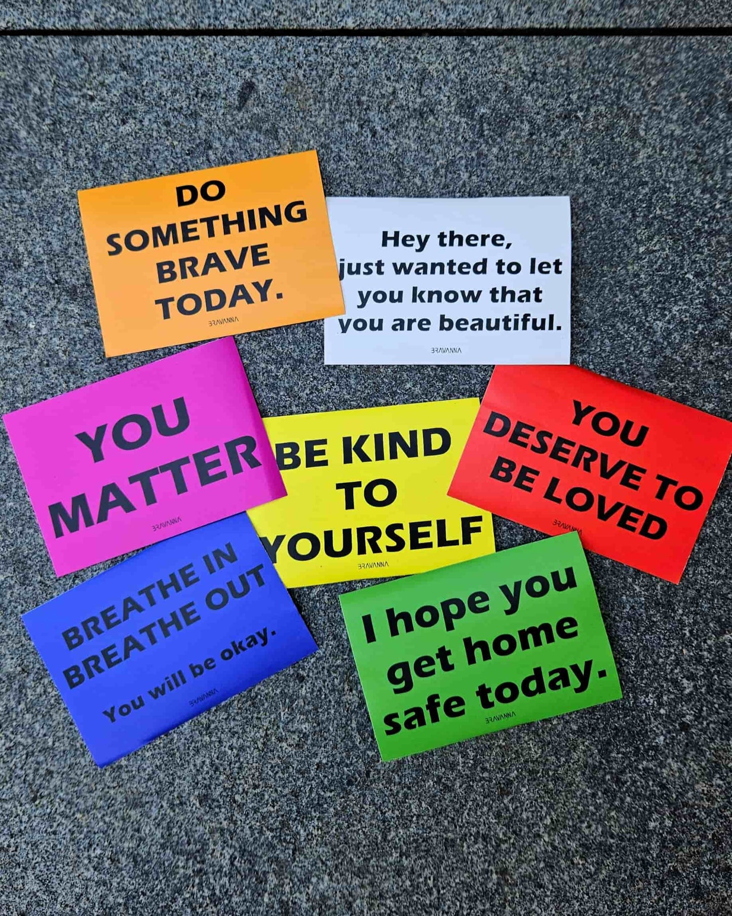 Stone floor with seven colorful rectangular stickers laying on it. The colors are orange, pink, beige, red, yellow, blue and green. Messages on the stickers include each of these phrases: You matter, do something brave today, you deserve to be loved, be kind to yourself, breathe in breathe out you will be okay, I hope you get home safe today, hey there just wanted to let you know that you are beautiful.