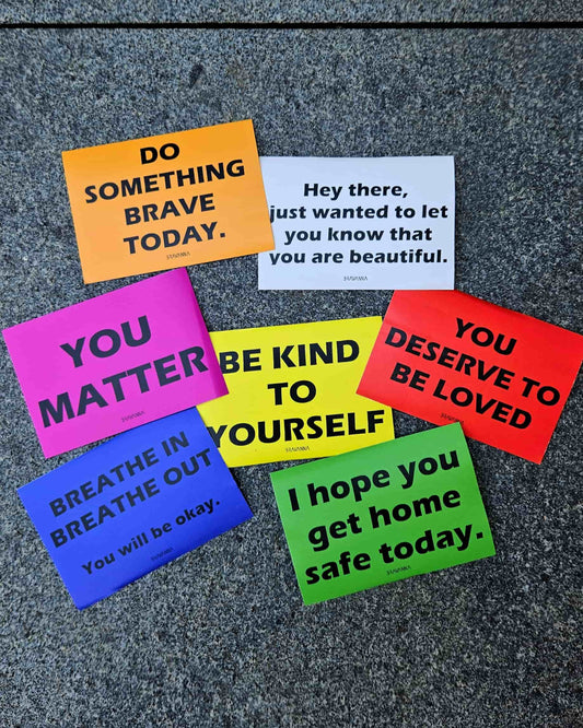 Stone floor with seven colorful rectangular stickers laying on it. The colors are orange, pink, beige, red, yellow, blue and green. Messages on the stickers include each of these phrases: You matter, do something brave today, you deserve to be loved, be kind to yourself, breathe in breathe out you will be okay, I hope you get home safe today, hey there just wanted to let you know that you are beautiful.